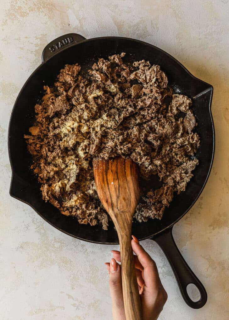 A hand stirring steak in a cast iron skillet on a tan counter.