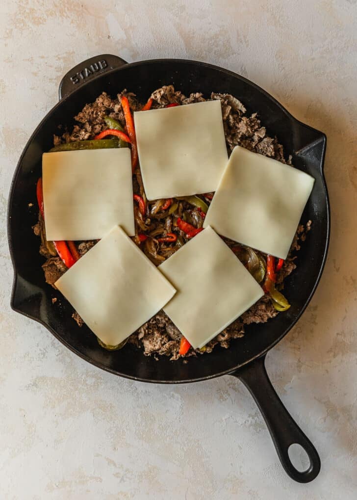 A cast iron skillet with meat, veggies, and cheese slices on a tan background.