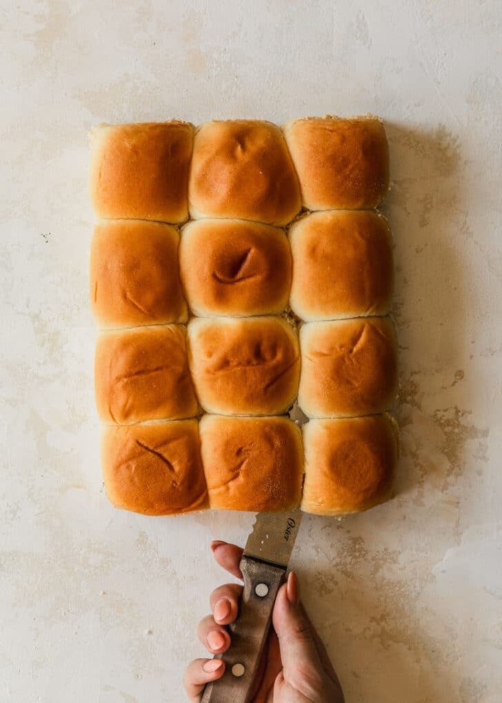 A hand using a knife to slice Hawaiian rolls in half on a tan backdrop.