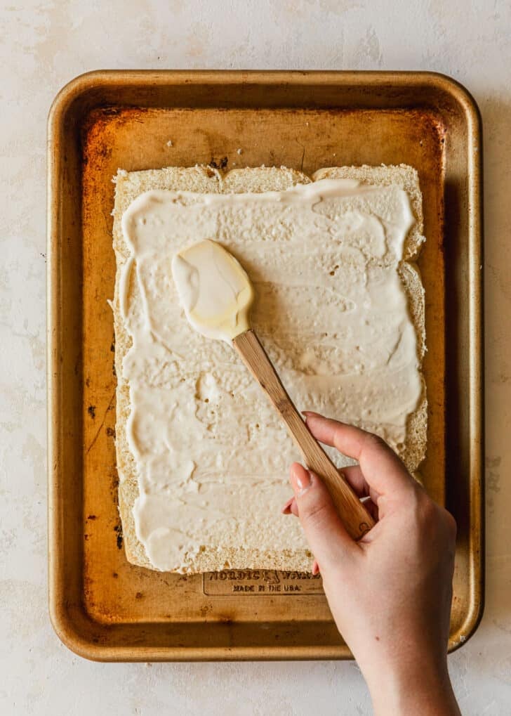 A hand using a spatula to spread mayo on Hawaiian rolls.