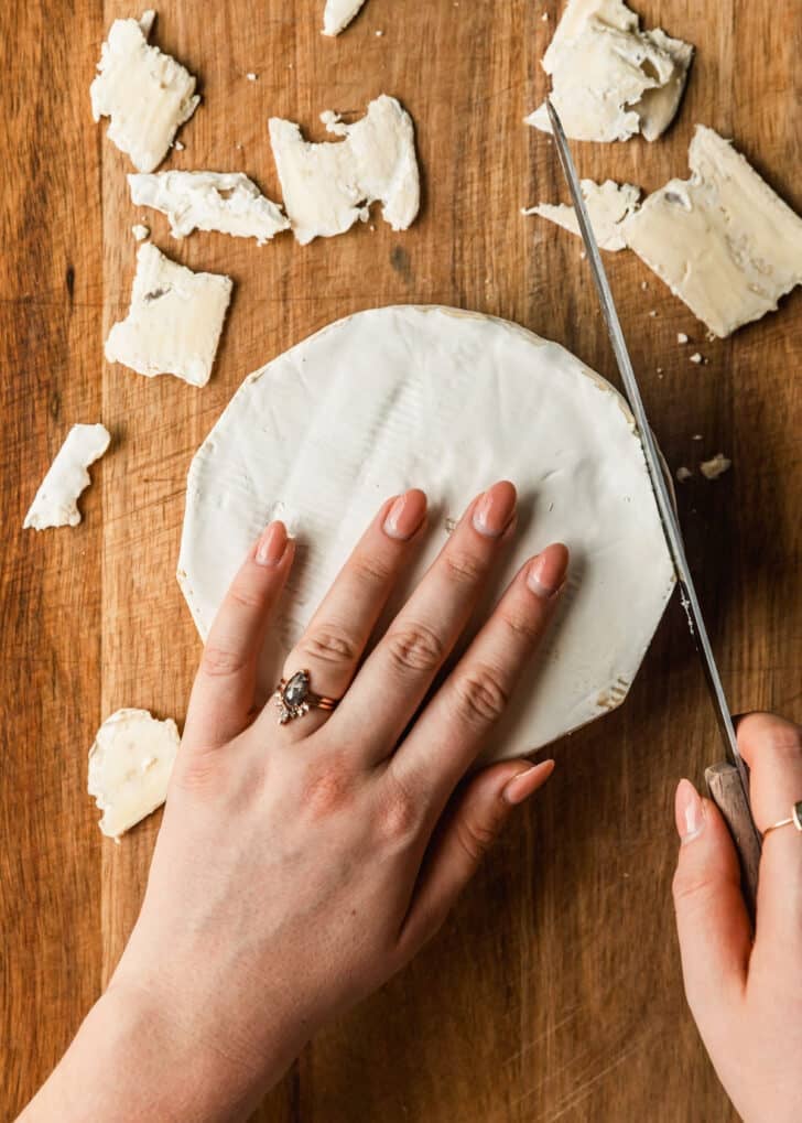 A hand slicing rind off of brie on a wood board.