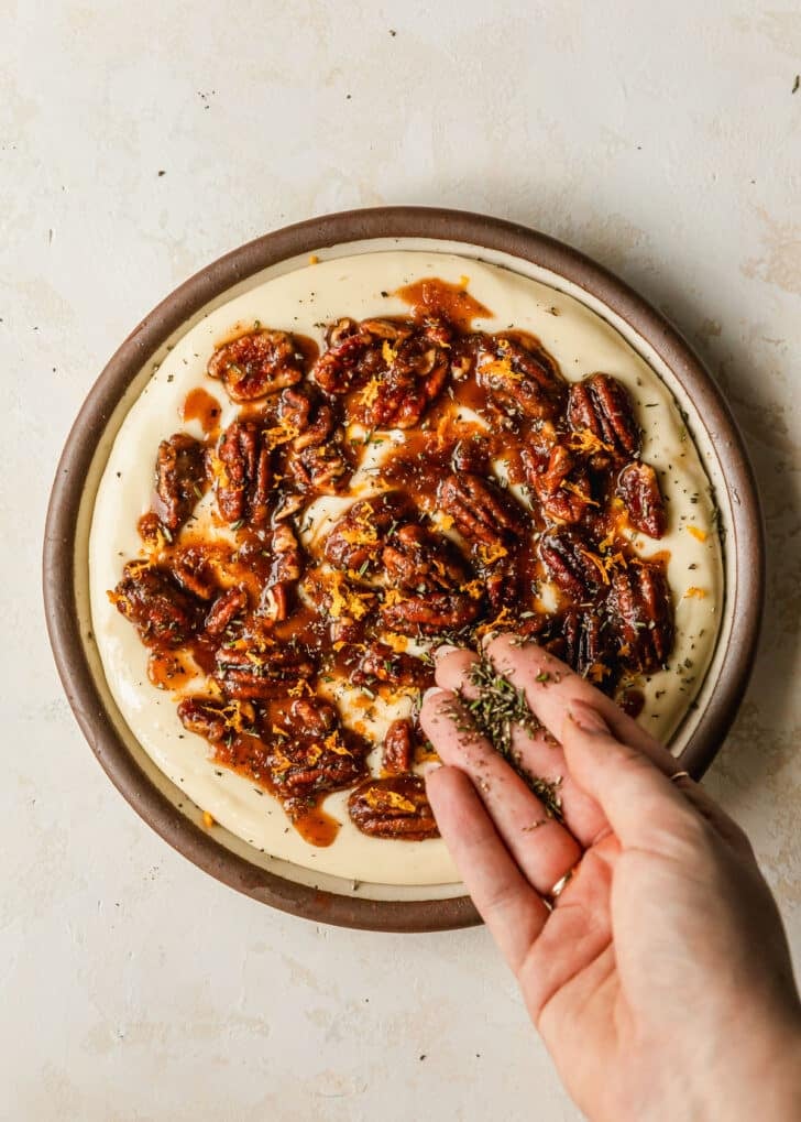 A hand sprinkling herbs over a brown plate of whipped brie cheese with pecans on a white counter.