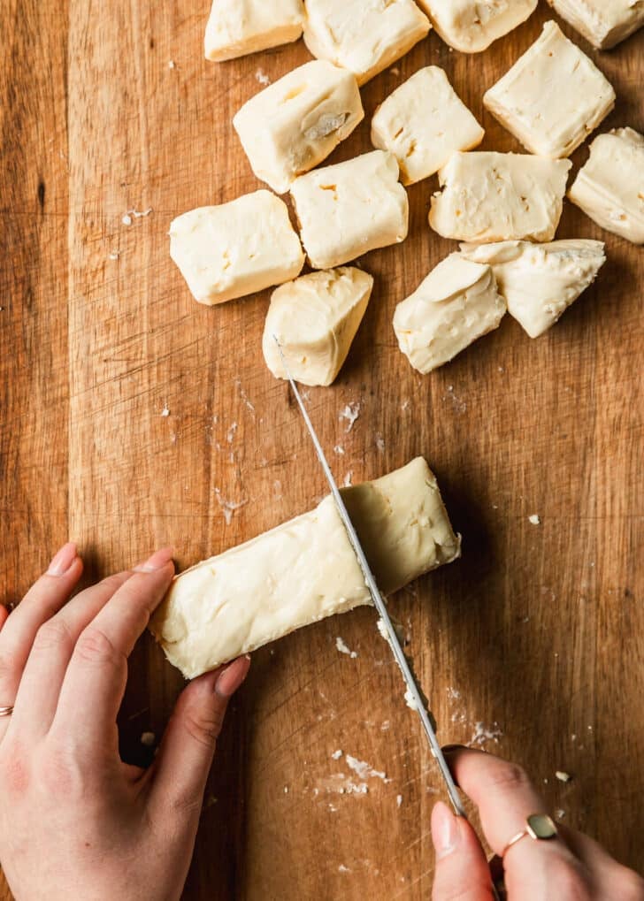 Hands slicing brie on a wood board.