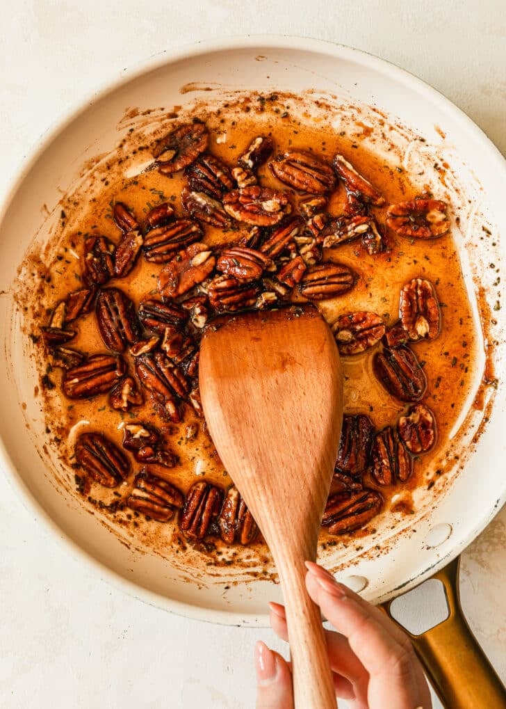 A hand stirring sticky pecans in a white pan on a white counter.