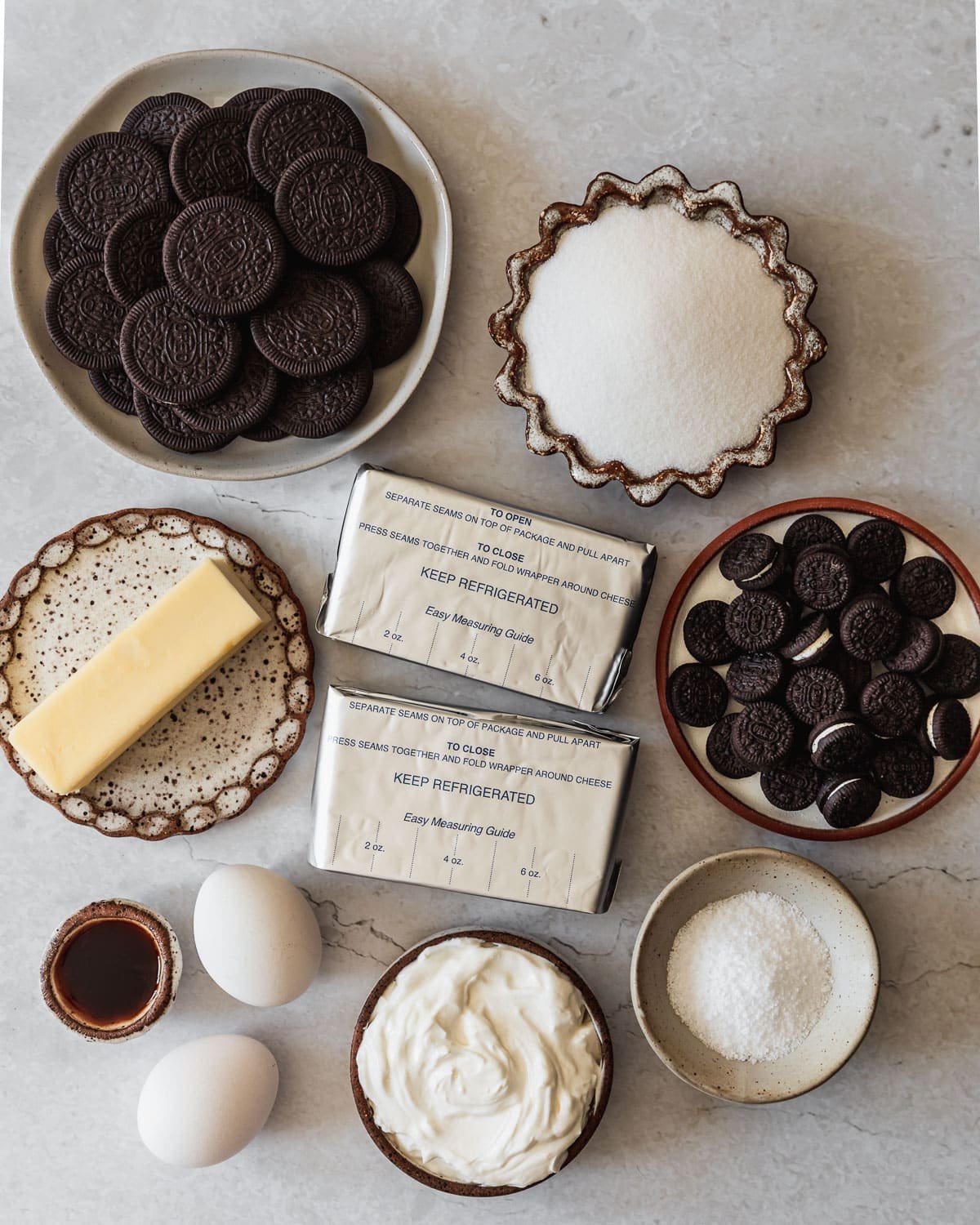 White and brown bowls of Oreos, sugar, butter, cream cheese, salt, sour cream, eggs, and vanilla on a marble counter.