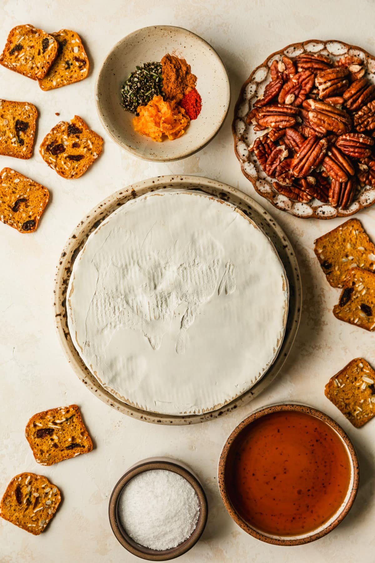 White and brown bowls of brie, pecans, orange zest, cinnamon, cayenne, rosemary, maple syrup, and salt on a tan counter.