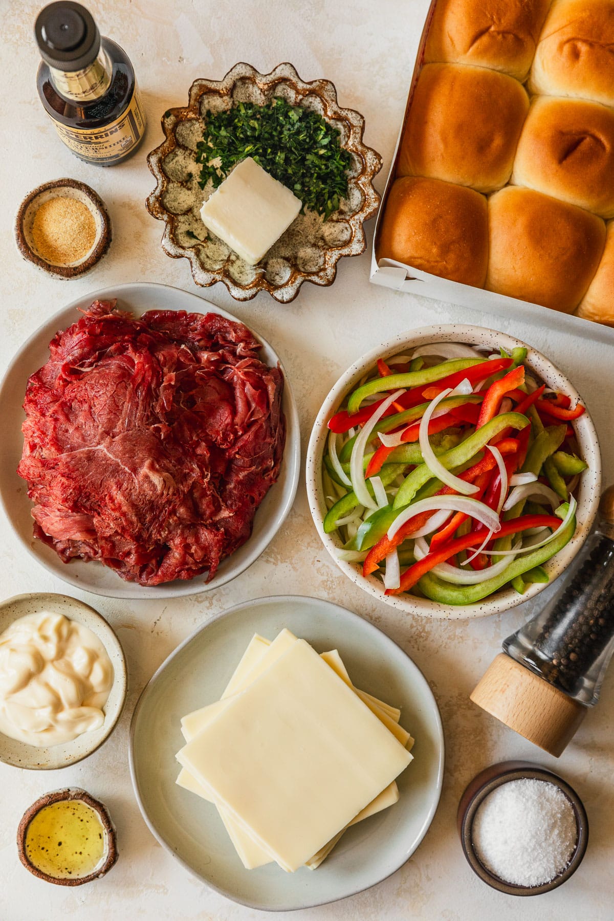 White and brown bowls of steak, peppers, onions, oil, salt, mayo, cheese, butter, parsley, and garlic powder on a tan counter next to rolls, Worcestershire sauce, and pepper.