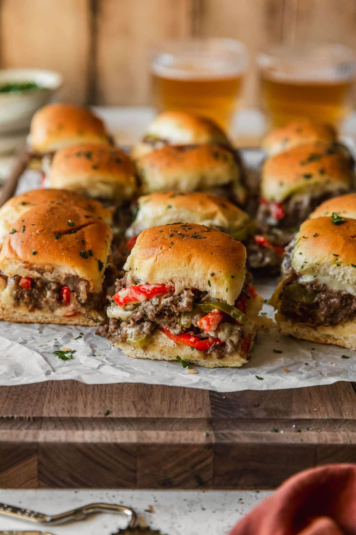 Philly cheesesteak sliders with Hawaiian rolls on a wood board next to glasses of beer and an orange linen on a wood backdrop.