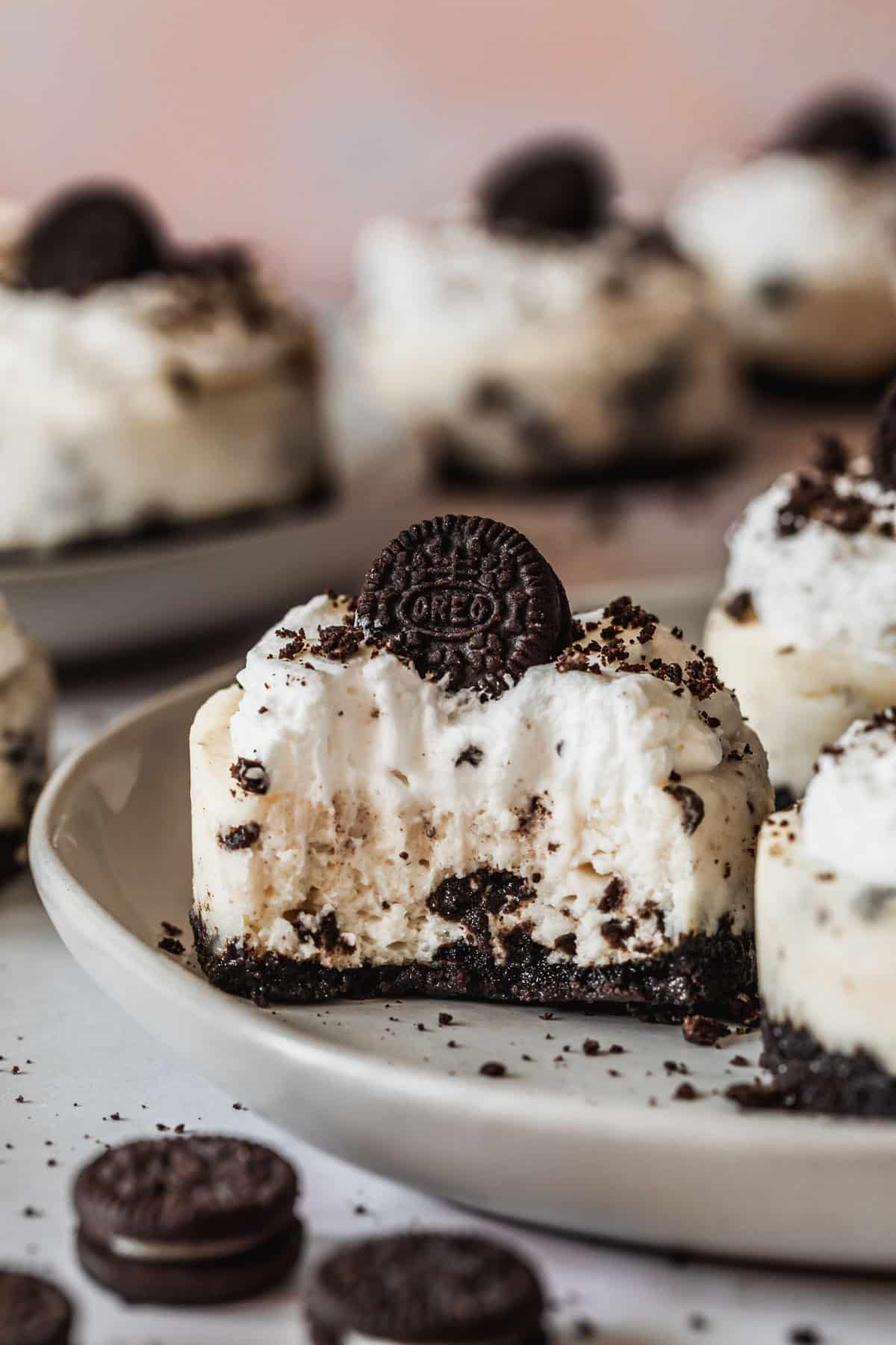 A mini Oreo cheesecake with a bite taken out of it on a white plate next to more mini Oreo cheesecakes and mini Oreos on a pink counter.