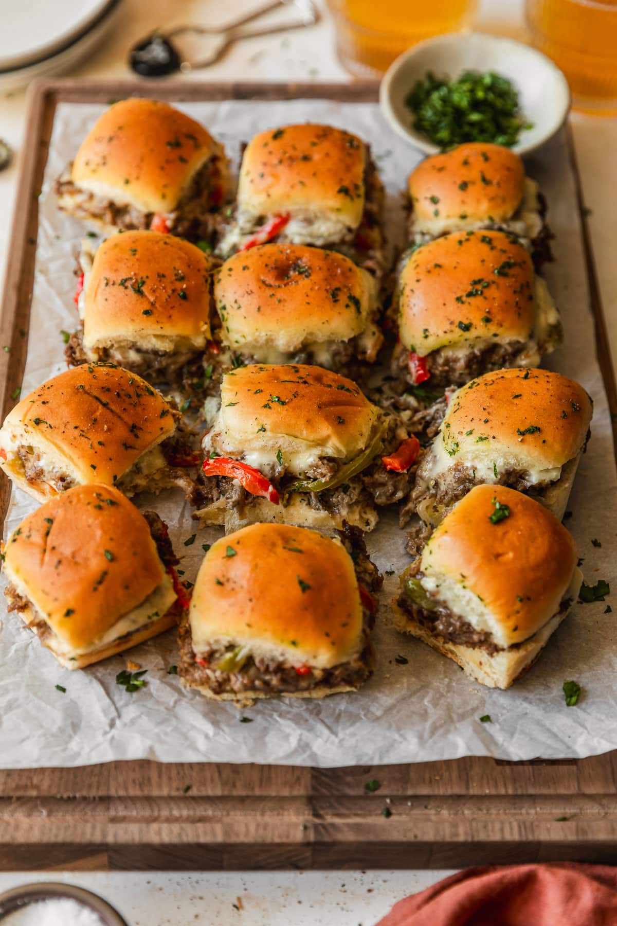 Rows of Philly cheesesteak sliders with Hawaiian rolls on a wood board next to glasses of beer, a white bowl of parsley, and an orange linen.