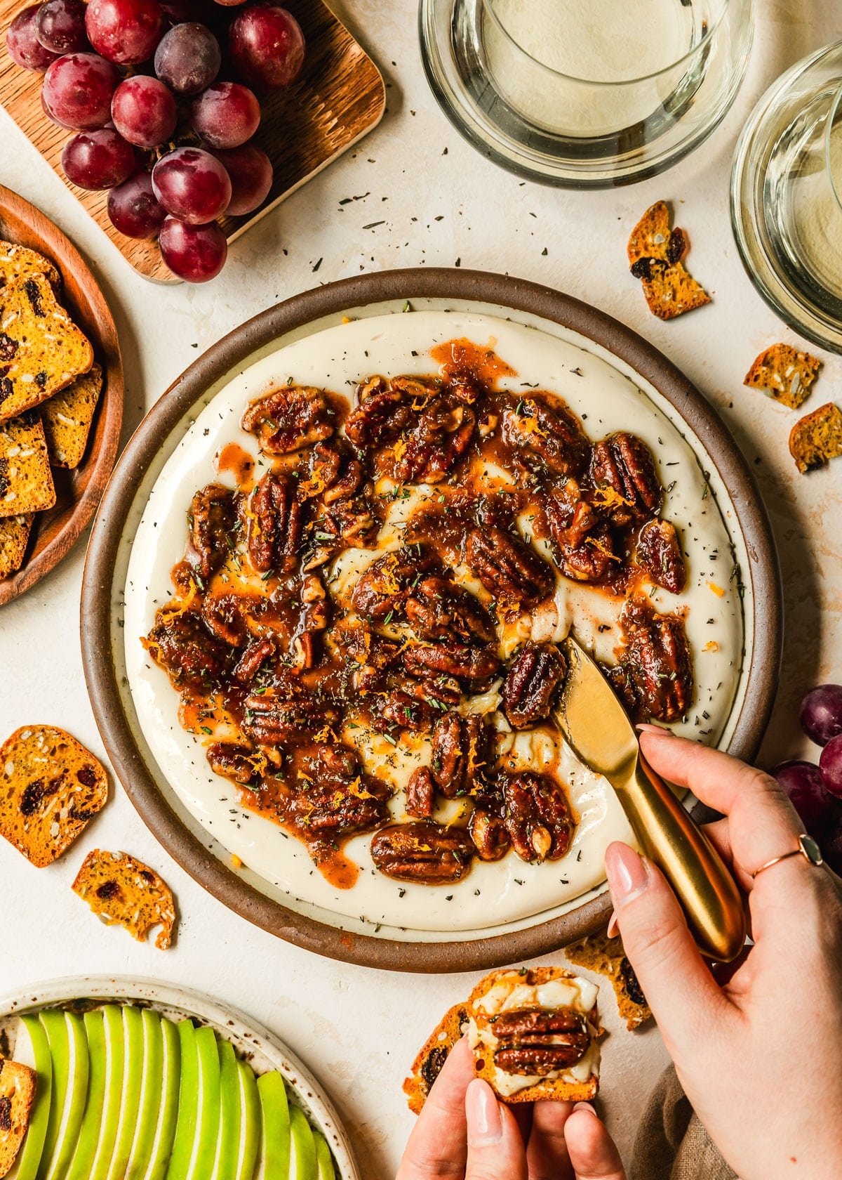 A hand using a gold cheese knife to scoop whipped brie cheese off of a stoneware plate next to glasses of white wine and brown plates of grapes, sliced green apples, and crackers with a tan background.