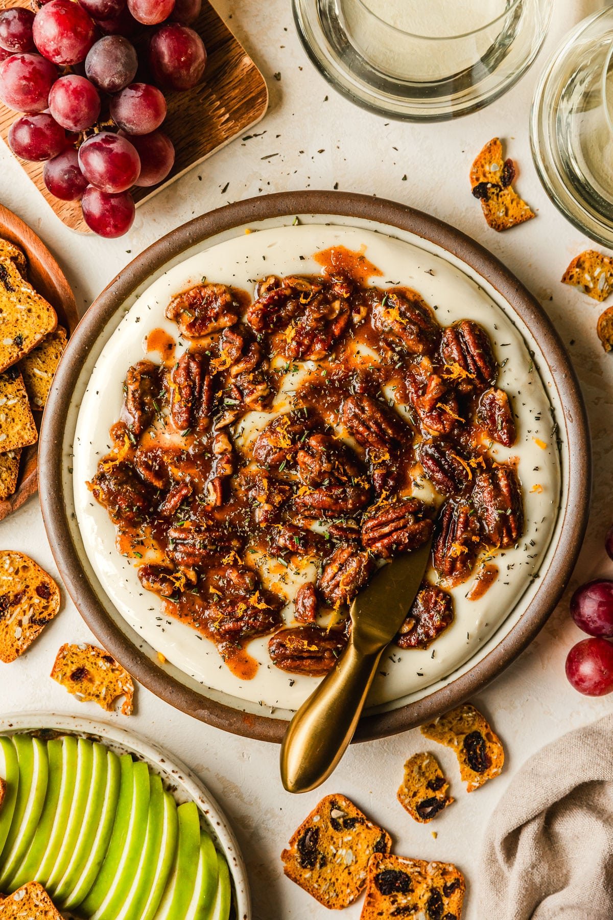 A brown plate of whipped brie cheese with sticky pecans on a tan counter next to glasses of white wine and brown plates of grapes, crackers, and green apple slices.