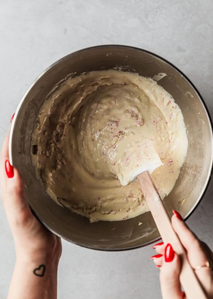 A hand using a rubber spatula to stir peppermint bark into mini cheesecakes filling on a white counter.