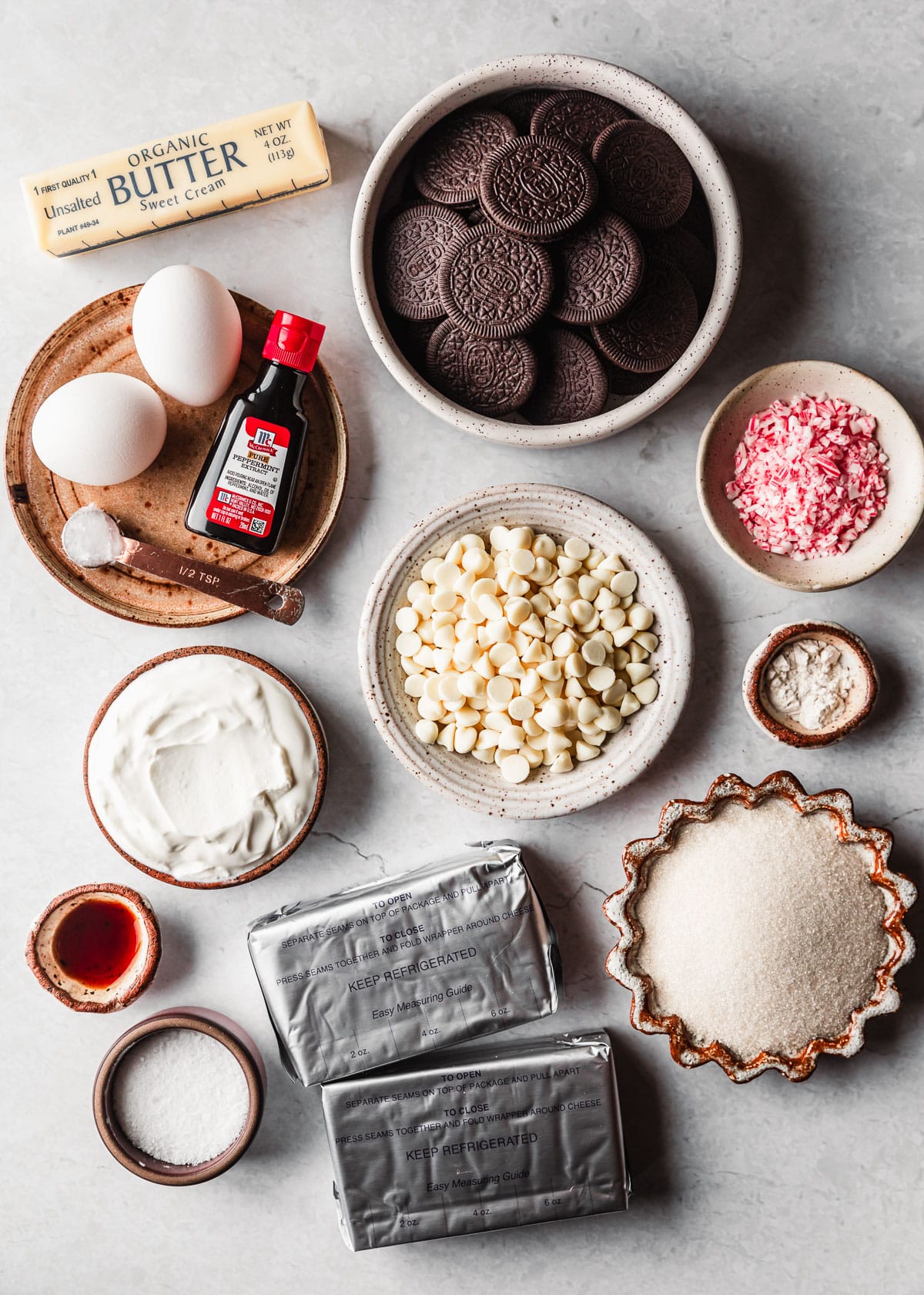 White and brown bowls of Oreos, peppermint candies, white chocolate, flour, eggs, peppermint extract, sour cream, butter, cream cheese, sugar, salt, and vanilla on a white counter.