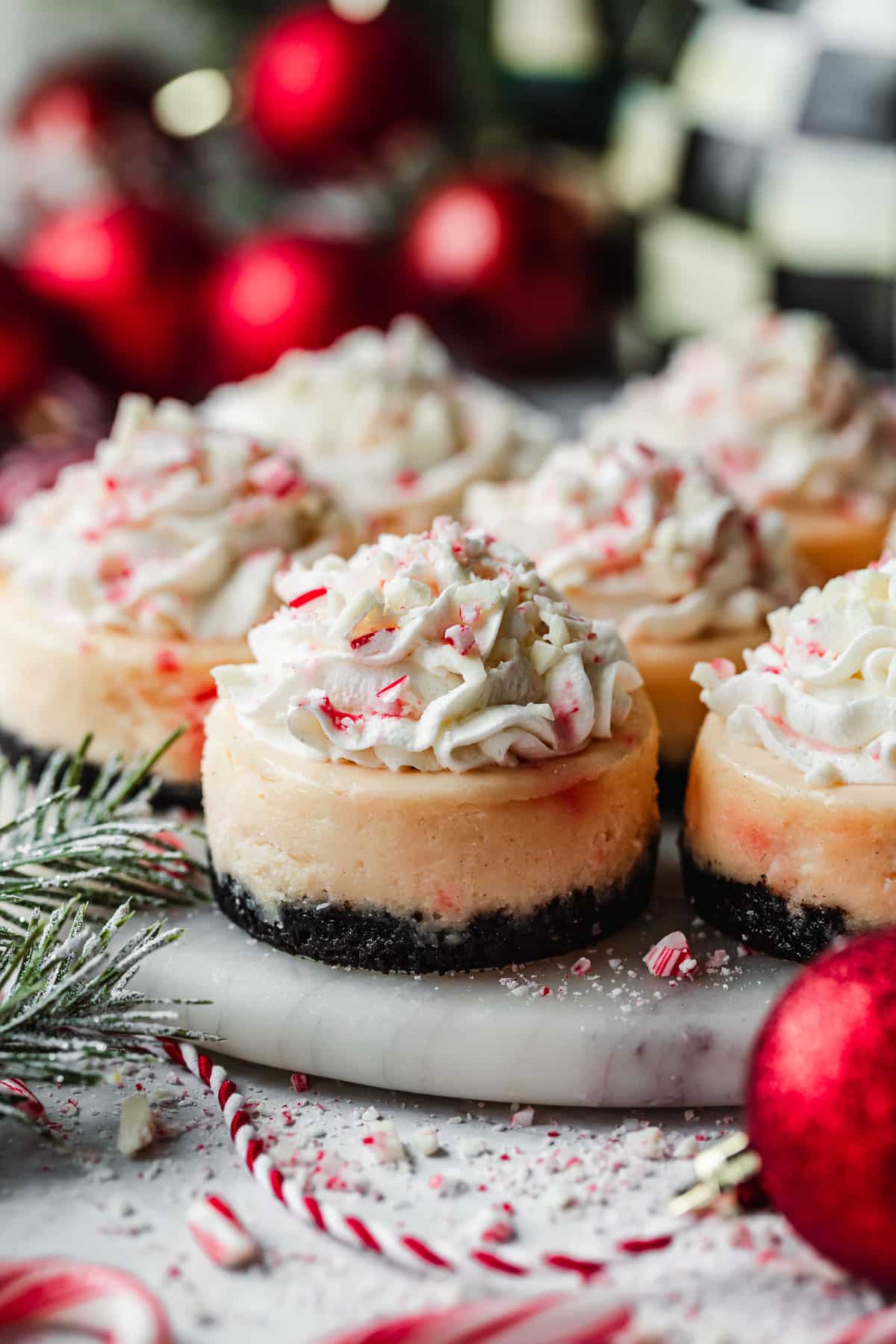 A marble platter with rows of mini peppermint bark cheesecakes next to red ornaments, garland, candy canes, and a checkered kettle with a white background.