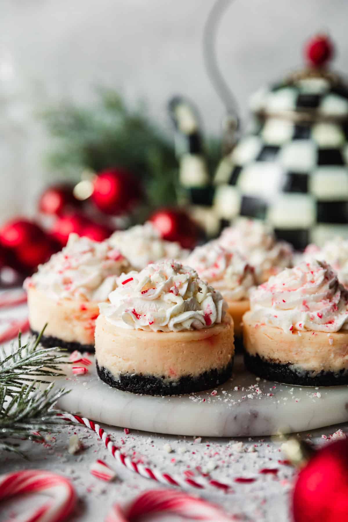 Rows of mini peppermint bark cheesecakes on a marble platter next to a checkered tea kettle, red ornaments, candy canes, and garland with a white background.