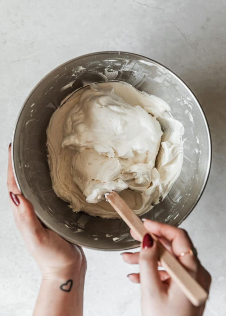 A hand using a rubber spatula to fold whipped cream into mini no-bake cheesecakes filling.