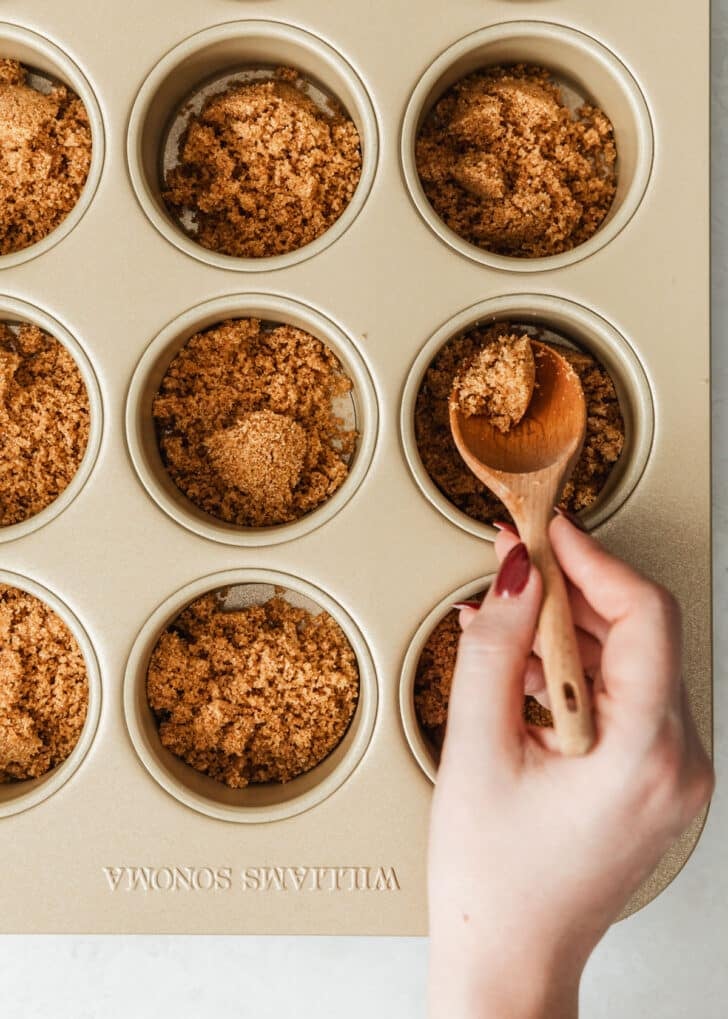 A hand spooning graham cracker crust into mini cheesecake pans.