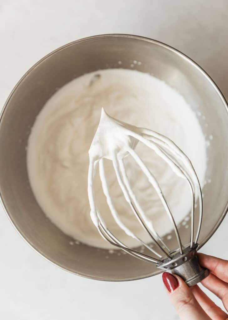 A hand holding a whisk with whipped cream over a silver bowl of whipped cream.