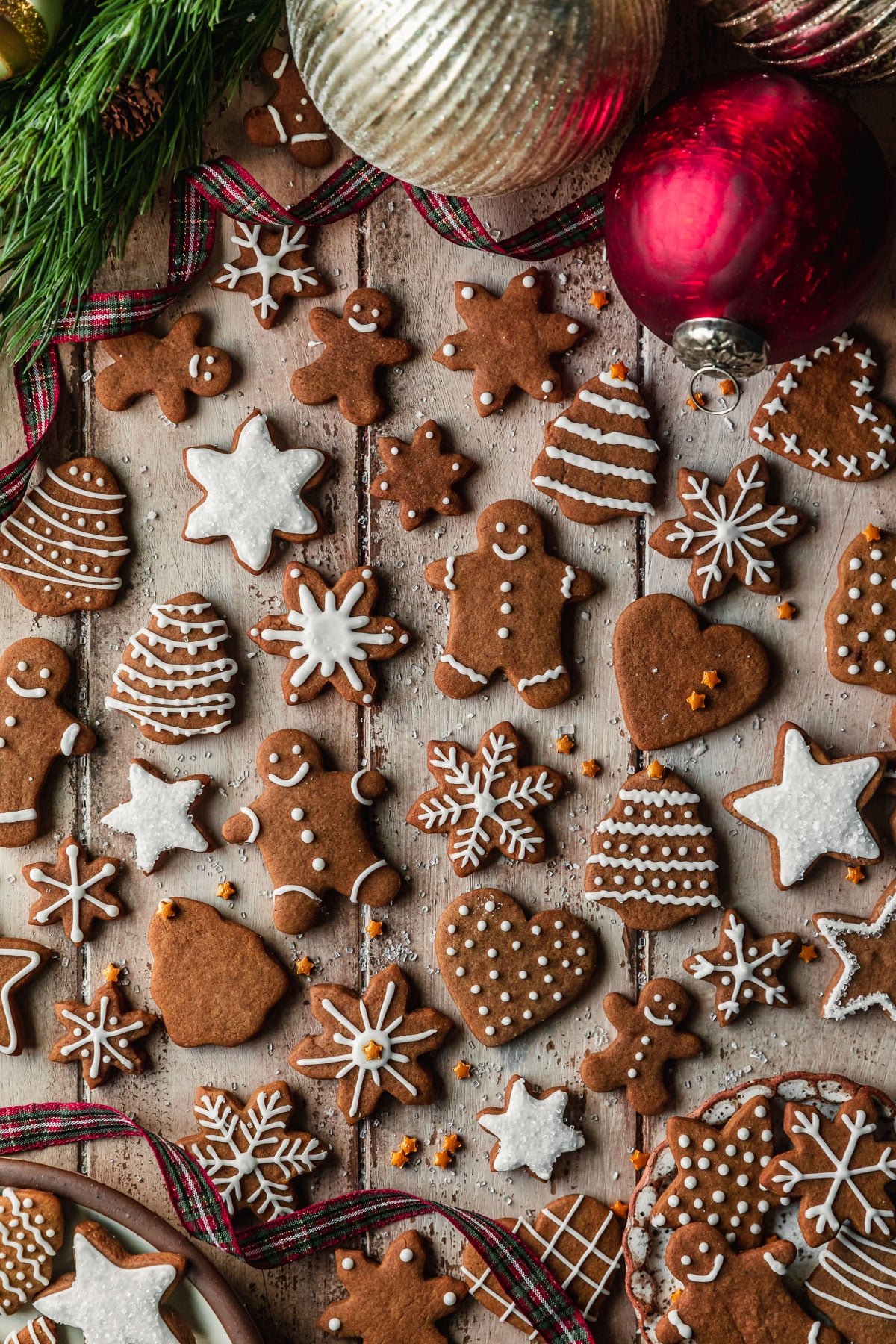 Rows of mini gingerbread cookies with maple icing on a wood table next to ornaments, garland, and red and green plaid ribbon.