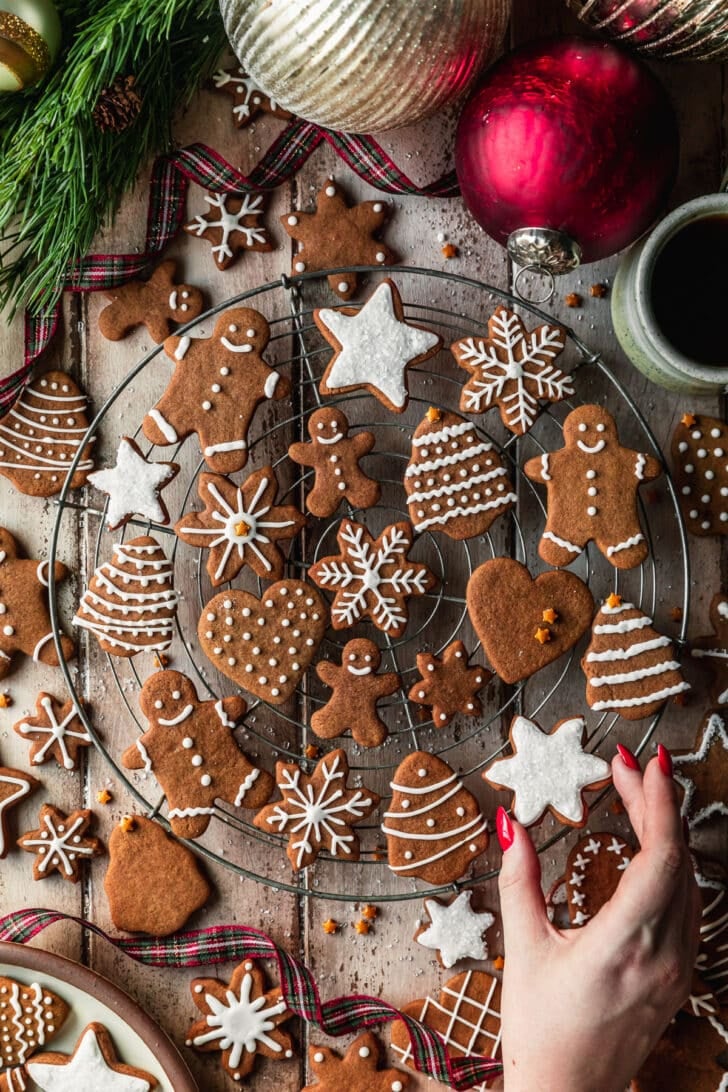 A hand grabbing a cookie off of a wire rack of mini gingerbread cookies next to plaid ribbon, garland, and ornaments with a wood background.