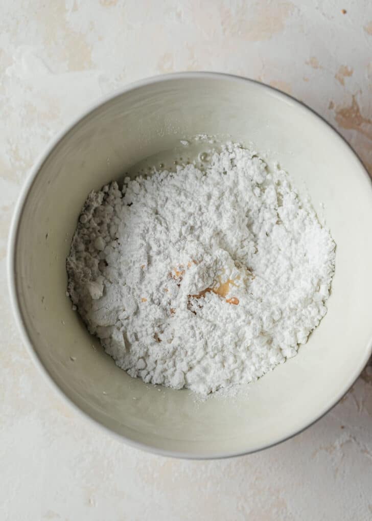 A white bowl with powdered sugar, milk, and maple extract on a tan counter.