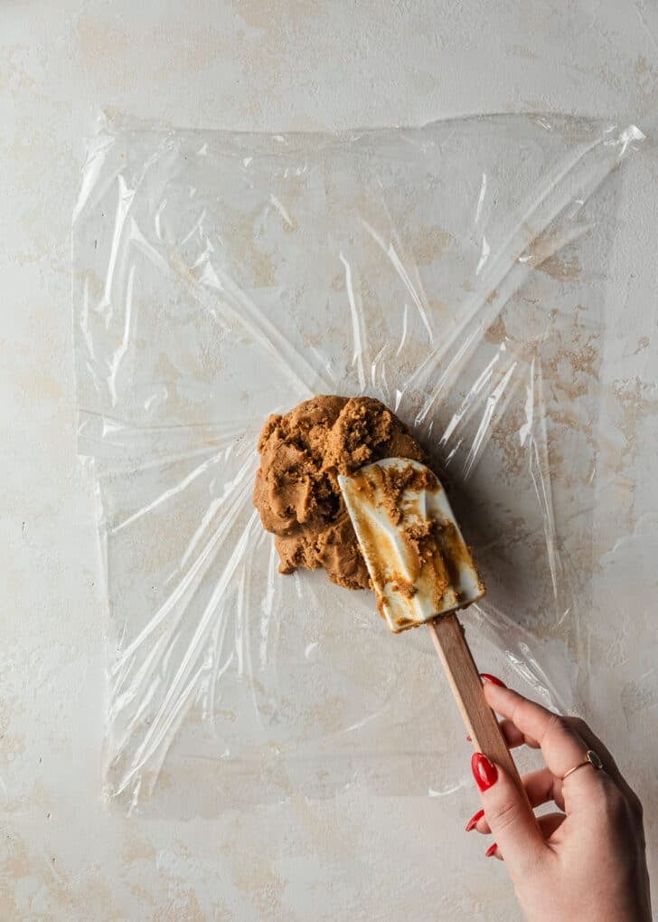 A hand using a rubber spatula to spoon gingerbread dough in the middle of plastic wrap on a tan counter.