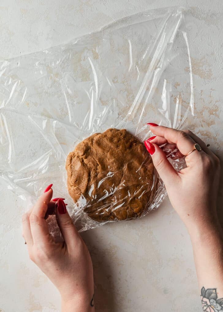 Hands wrapping cookie dough in plastic wrap on a beige counter.