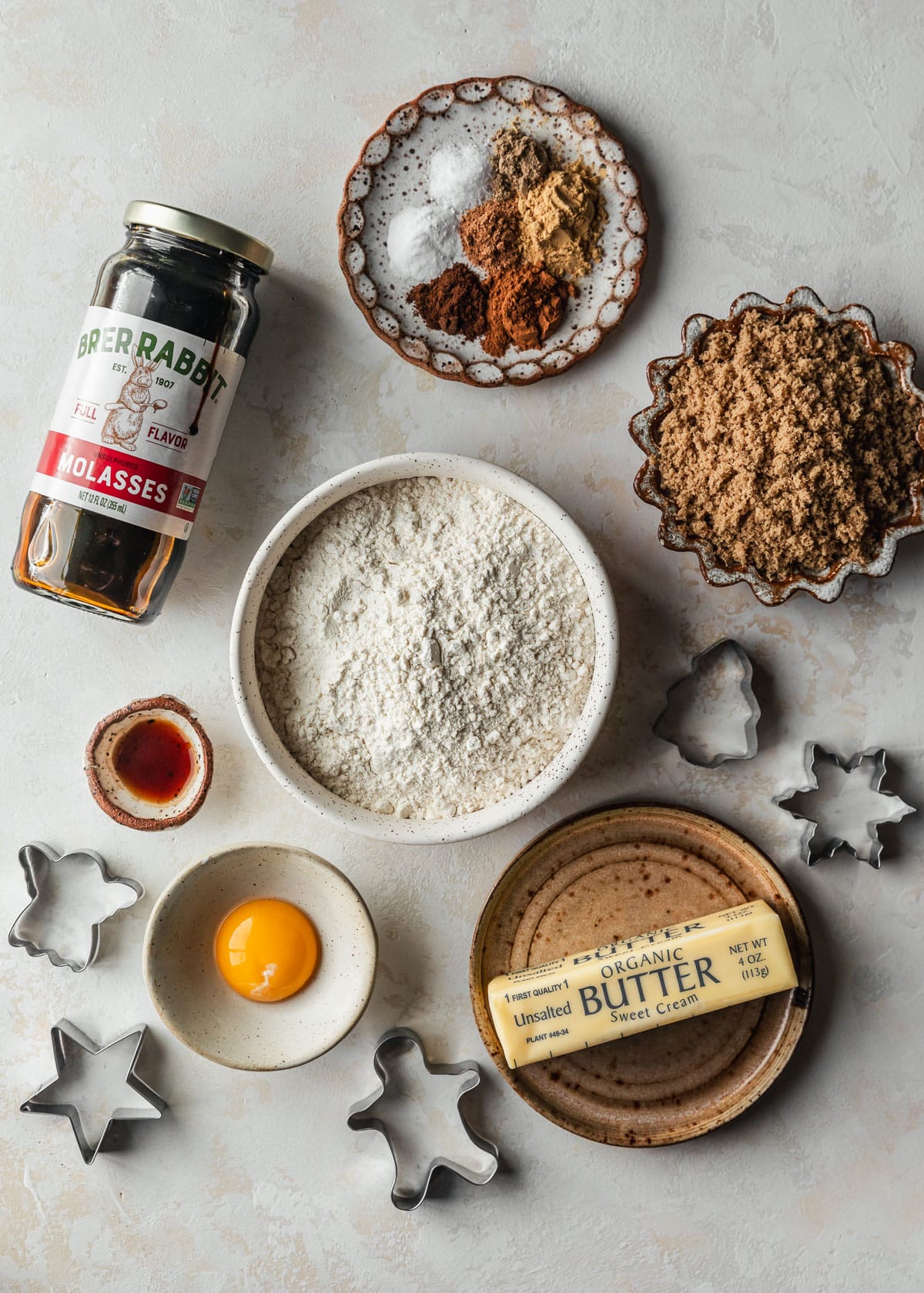 White bowls of spices, brown sugar, flour, vanilla, butter, and an egg on a tan counter next to cookie cutters and a jar of molasses.