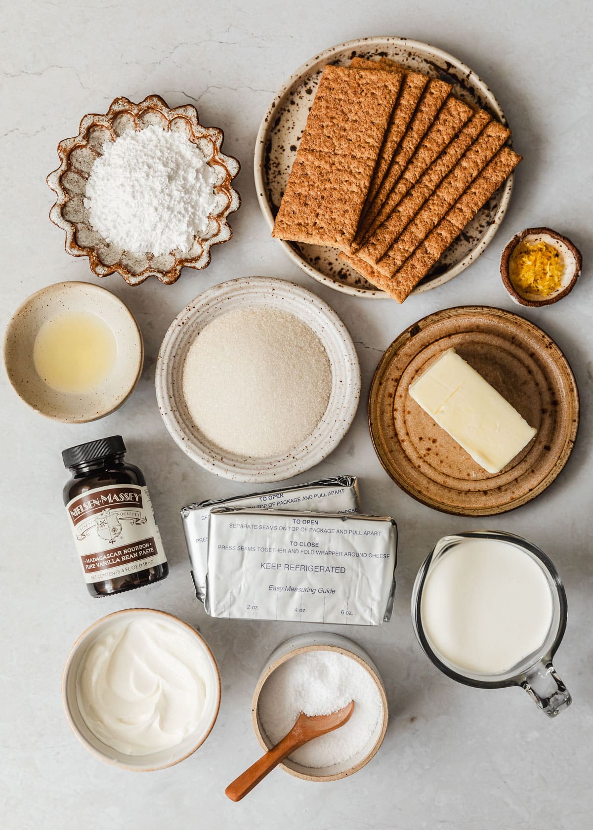 White and brown bowls of graham crackers, powdered sugar, lemon zest and juice, sugar, butter, cream, sour cream, and salt next to vanilla and cream cheese on a white counter.