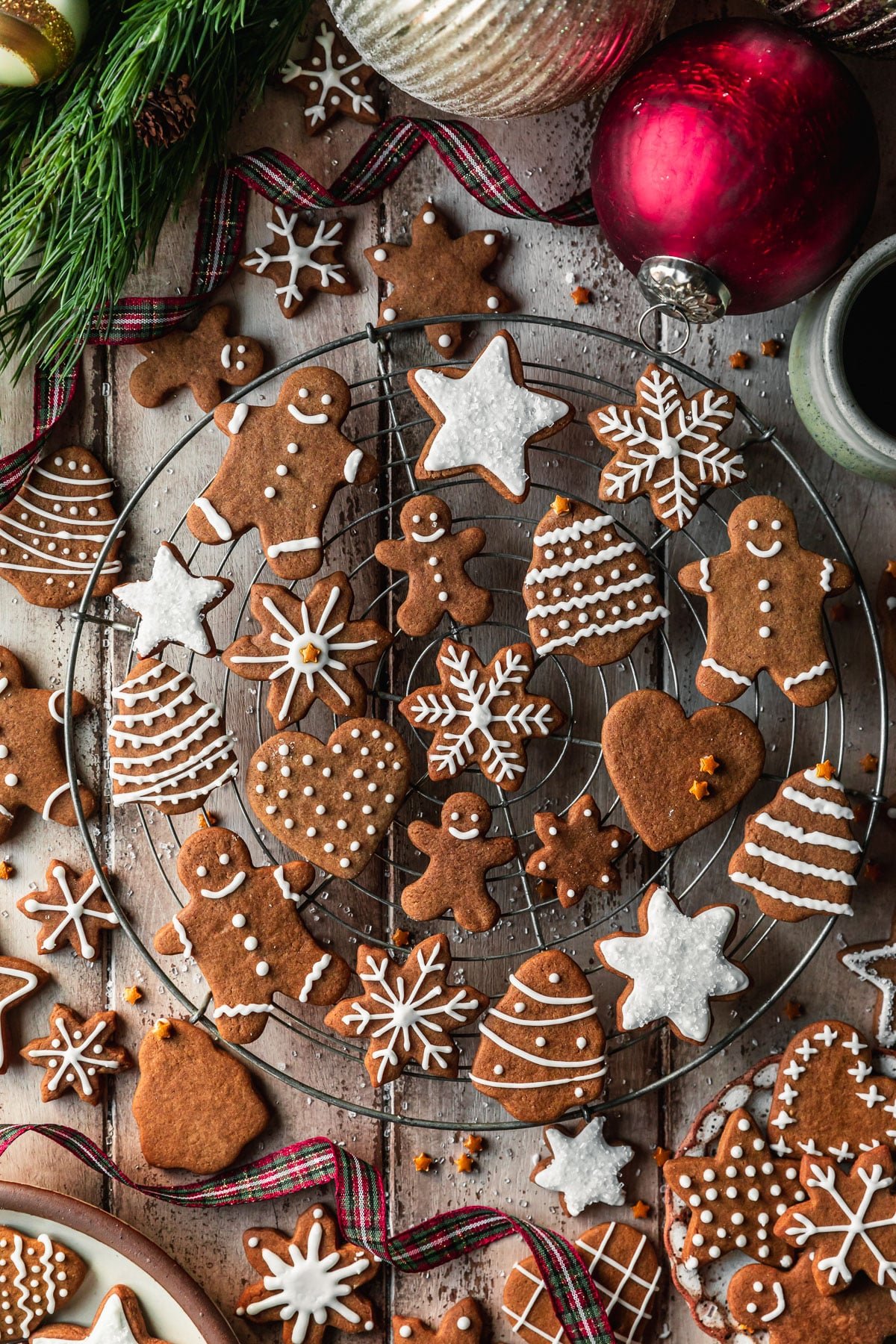 A wire rack with mini gingerbread cookies on a wood table next to plaid Christmas ribbon, ornaments, and garland.