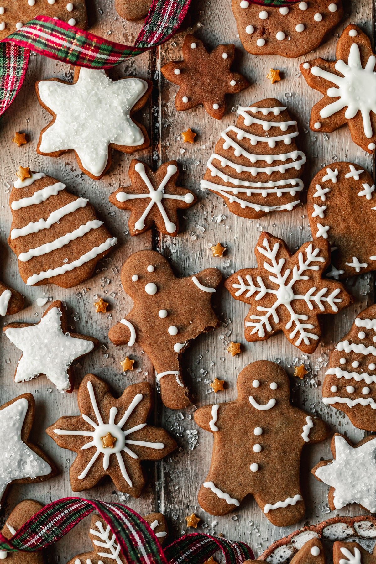 Rows of mini gingerbread cookies on a wood table next to red and green plaid ribbon and gold star sprinkles.