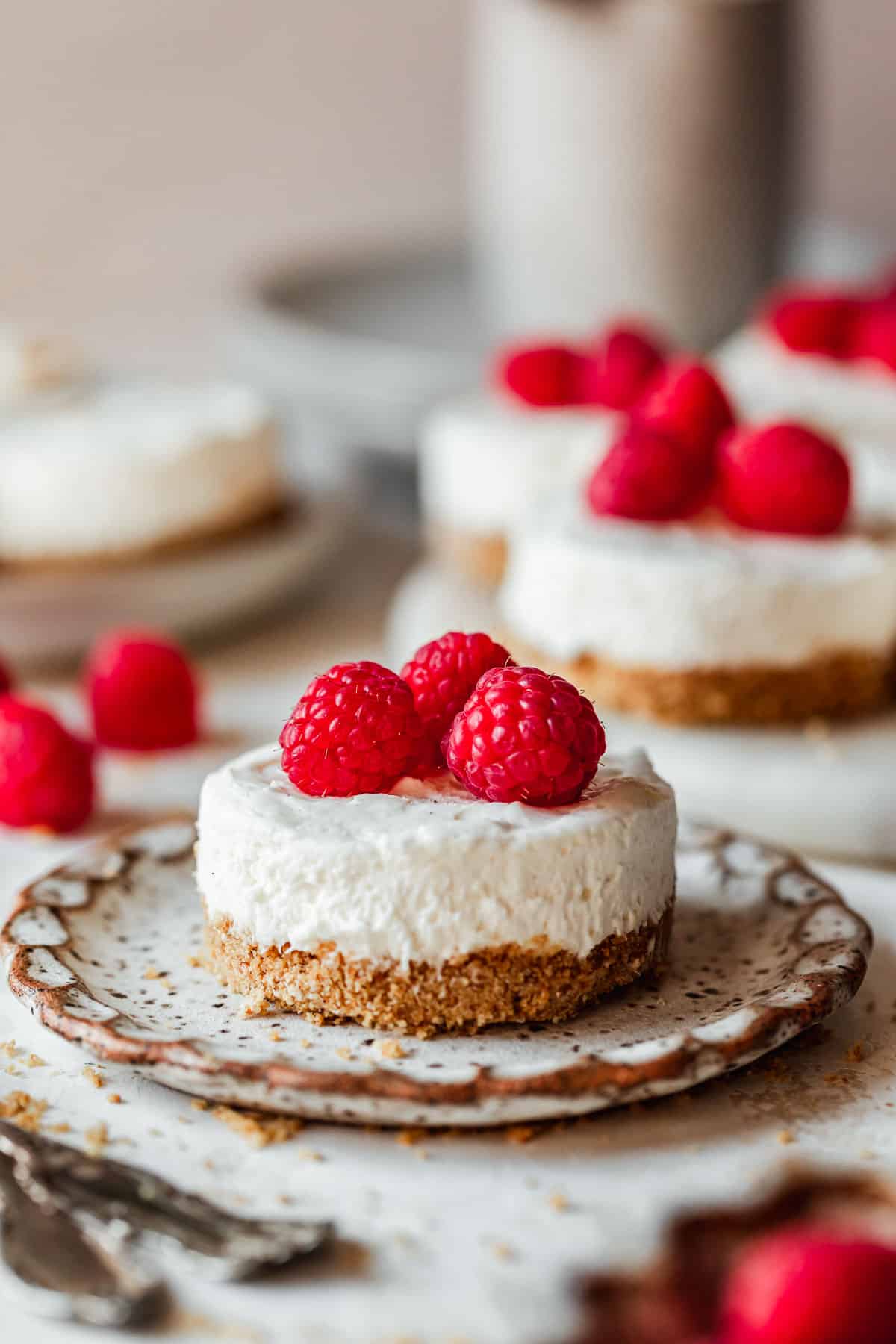 A cheesecake with raspberries on a white plate next to a white platter of mini no bake cheesecakes, raspberries, and a white carafe on a pink backdrop.