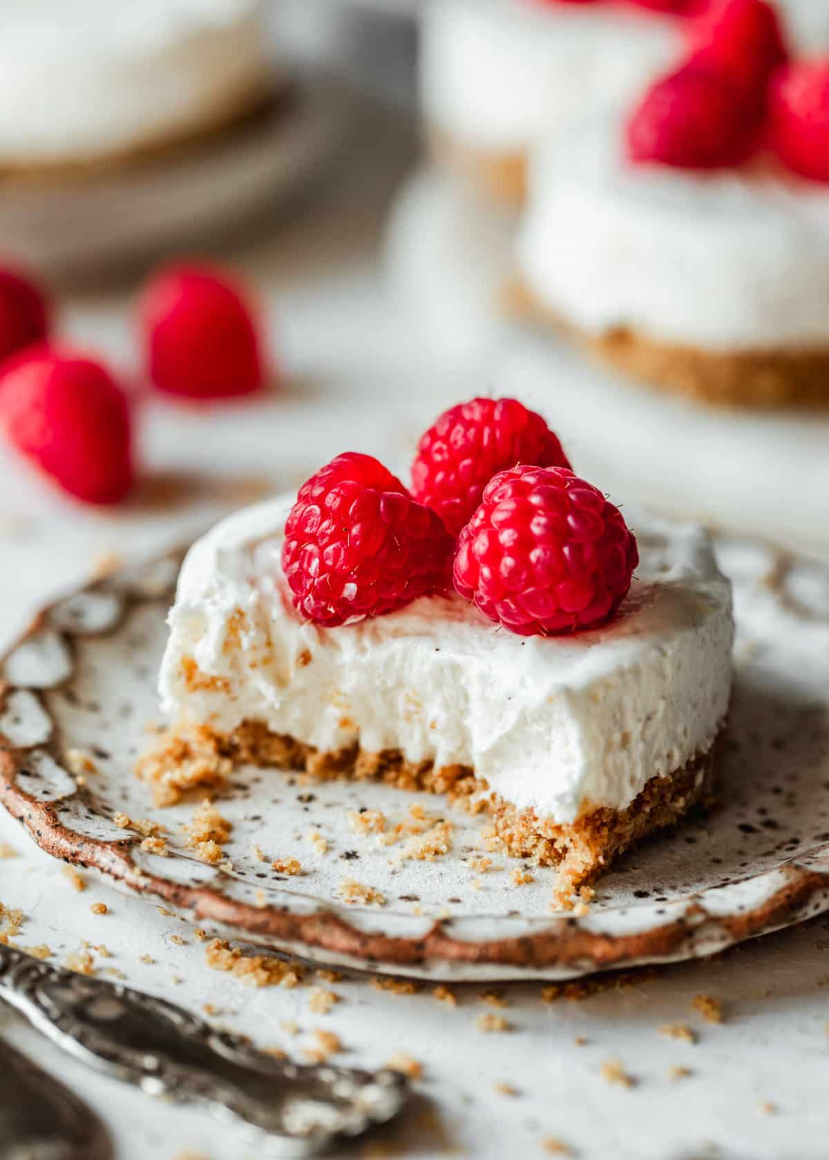 A white plate with a cheesecake with a bite taken out of it on a white counter next to a platter of mini no bake cheesecakes and raspberries.