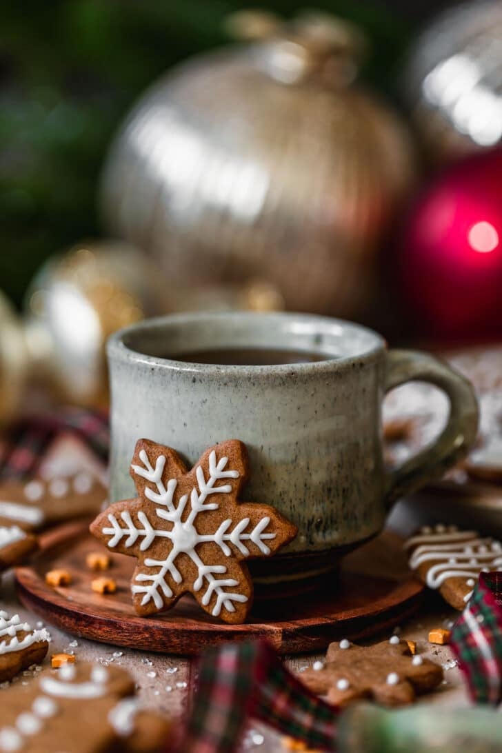 A snowflake mini gingerbread cookie leaning against a grey mug of coffee on a wood table next to ornaments, garland, and plaid ribbon.