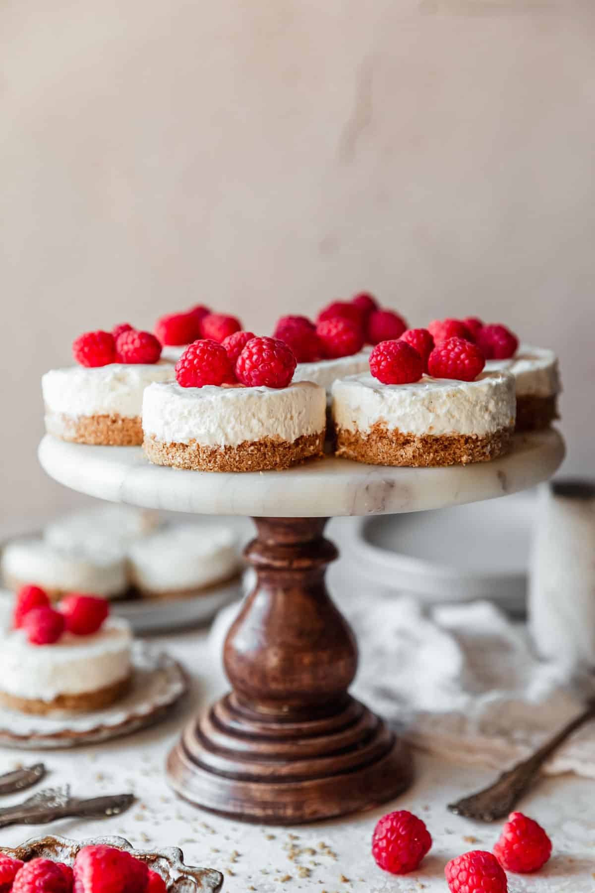 A marble cake stand with no bake mini cheesecakes on a pink backdrop next to a brown bowl of raspberries, white plates, and a white linen.