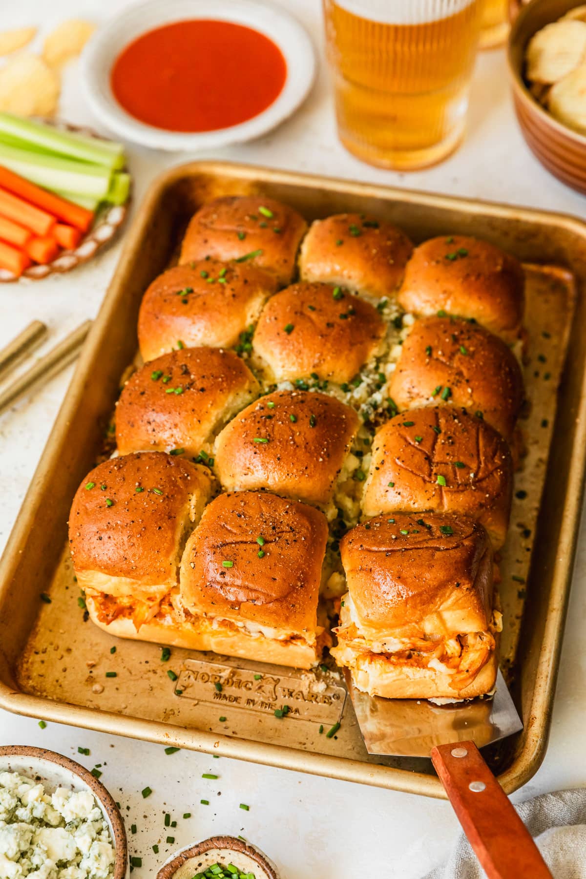 A gold sheet pan of buffalo chicken sliders on a tan counter next to glasses of beer, a brown bowl of chips, and white bowls of buffalo sauce, carrots, celery, and blue cheese.