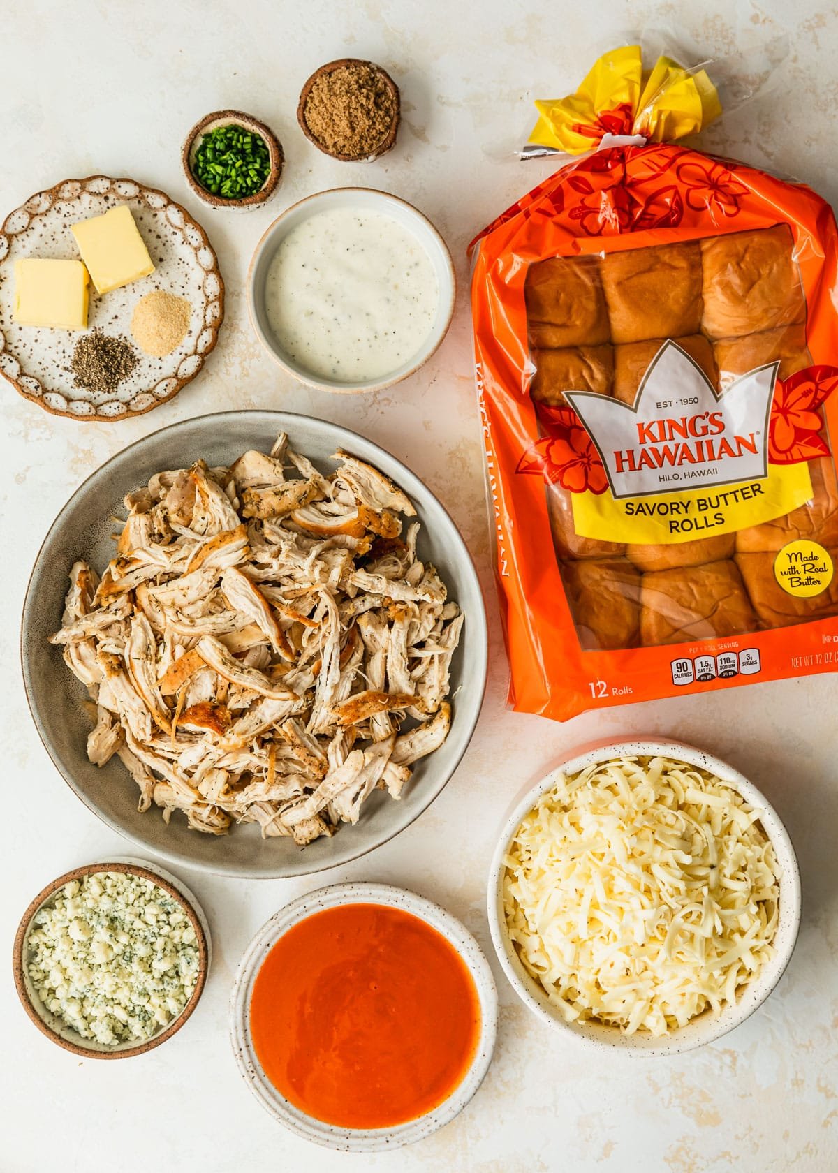 White and brown bowls of shredded chicken, brown sugar, chives, butter, spices, ranch, hot sauce, blue cheese, and pepper jack cheese next to a pack of Hawaiian rolls on a tan counter.