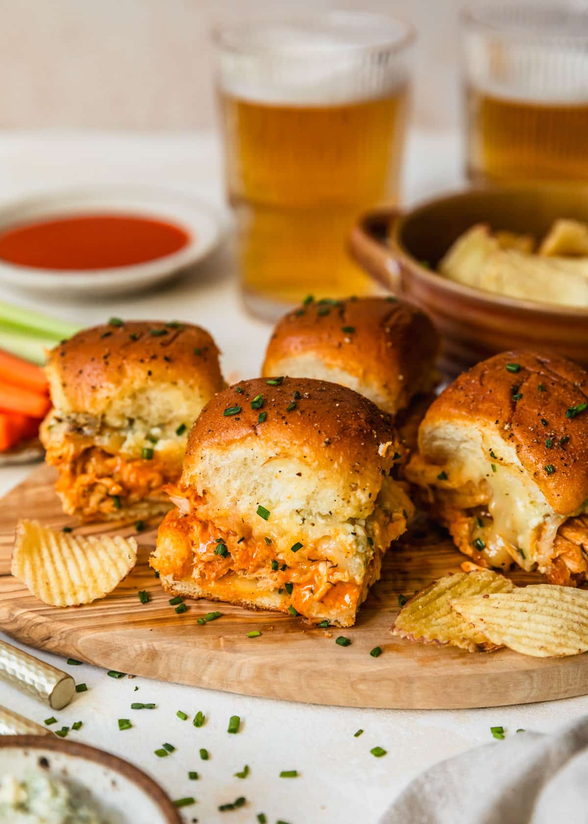 Buffalo chicken sliders on a wood board next to a brown bowl of potato chips, white bowls of buffalo sauce, carrots, celery, and blue cheese, and glasses of beer with a tan background.