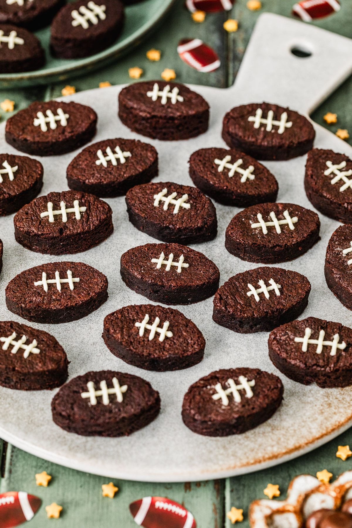 A grey tray of mini football brownies on a green table next to star sprinkles, football confetti, and a green plate of brownies.