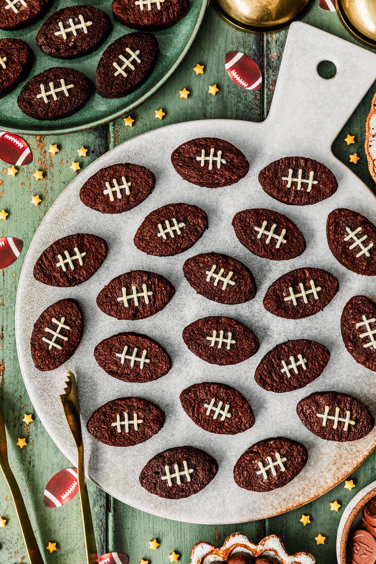 A grey platter of mini football brownies on a green wood table next to a green plate of brownies, white bowls of chocolates, star sprinkles, and football confetti.