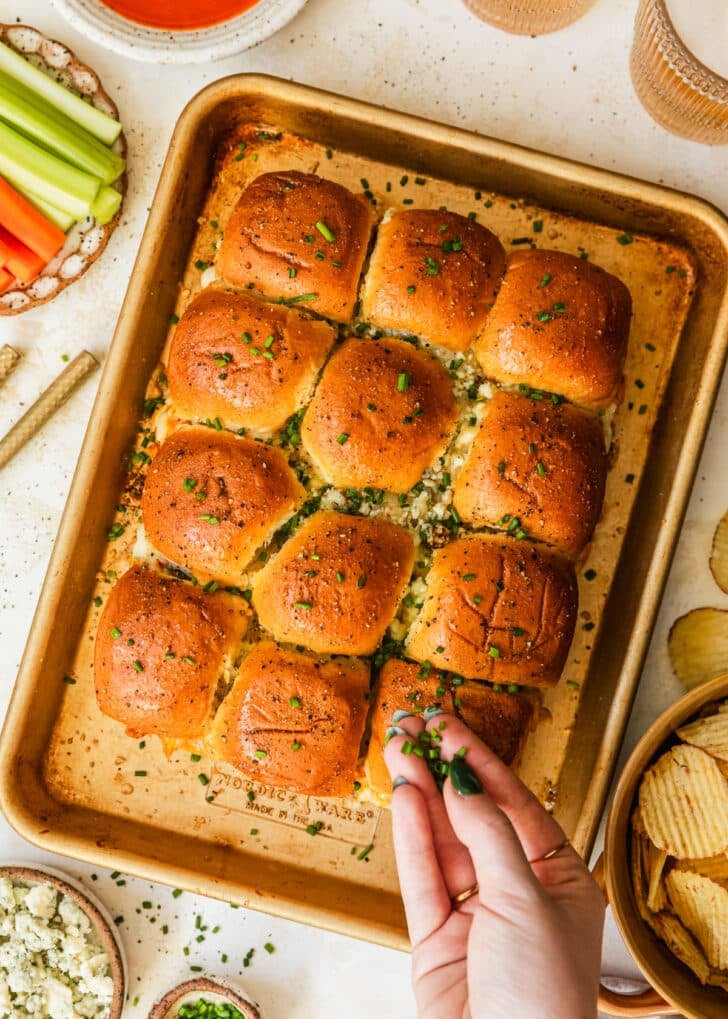 A hand sprinkling chives over a gold sheet pan of buffalo chicken sliders on a tan table next to glasses of beer, a white plate of carrots and celery, and a brown bowl of potato chips.