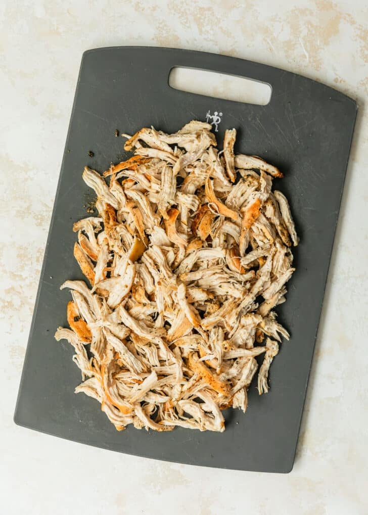 A grey cutting board with shredded chicken on a beige backdrop.