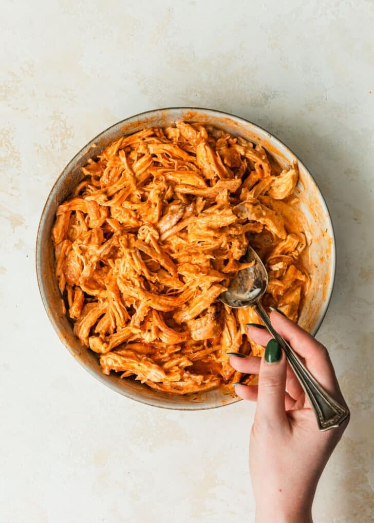 A hand using a spoon to mix buffalo chicken filling in a grey bowl on a beige backdrop.