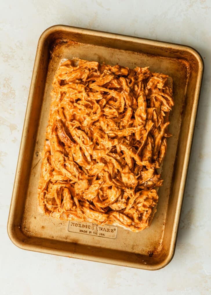 A sheet pan with Hawaiian rolls and buffalo chicken on a tan counter.