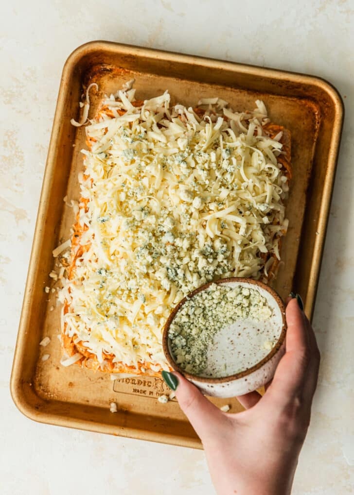 A hand sprinkling blue cheese over buffalo chicken sliders on a gold sheet pan.