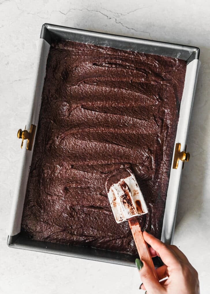 A hand using a rubber spatula to smooth brownie batter in a pan.