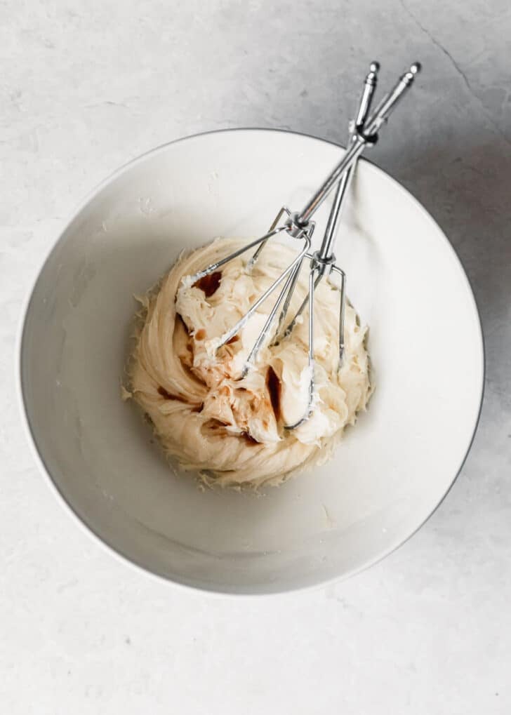 A white bowl of frosting, vanilla, and beaters on a white counter.