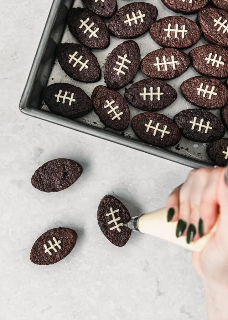 A hand using a piping bag to pipe laces on mini football brownies.