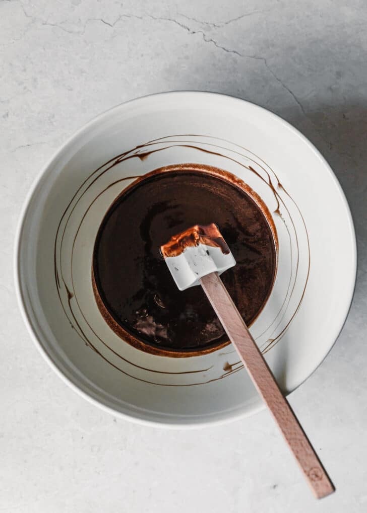 A white bowl of melted chocolate with a rubber spatula on a white counter.